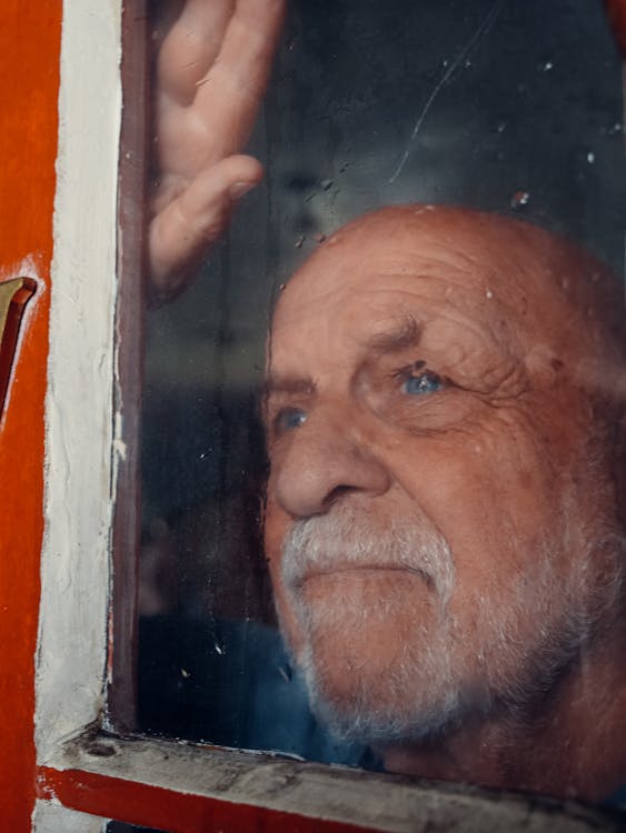 An elderly man looking through a glass window in contemplation